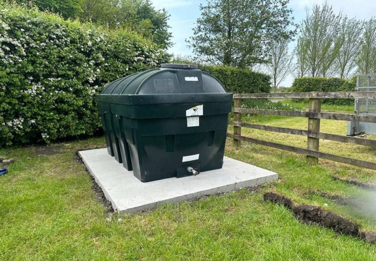 Green oil tank on a concrete base in a grassy yard beside hedges and a wooden fence
